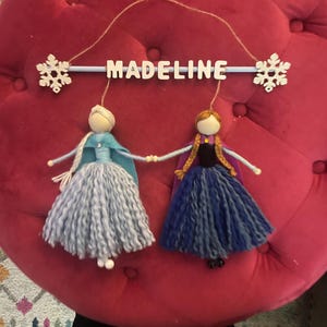 May include: A wooden mobile with the name "Madeline" spelled out in white letters. Two felt dolls, one in blue and one in purple, hang from the mobile. The dolls are dressed as Elsa and Anna from the movie Frozen.