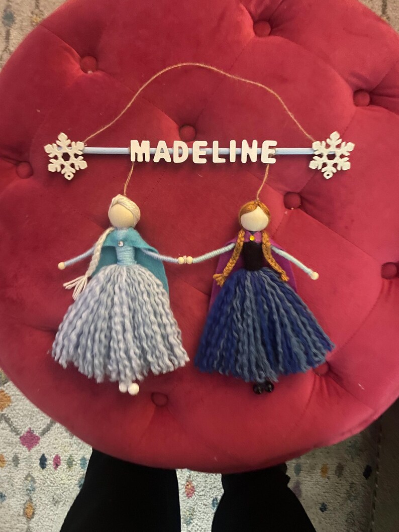 May include: A wooden mobile with the name "Madeline" spelled out in white letters. Two felt dolls, one in blue and one in purple, hang from the mobile. The dolls are dressed as Elsa and Anna from the movie Frozen.