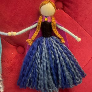 May include: A handmade felt doll with a wooden head, brown hair, and a blue and purple dress. The doll is wearing a purple cape and has a yellow button on her dress.