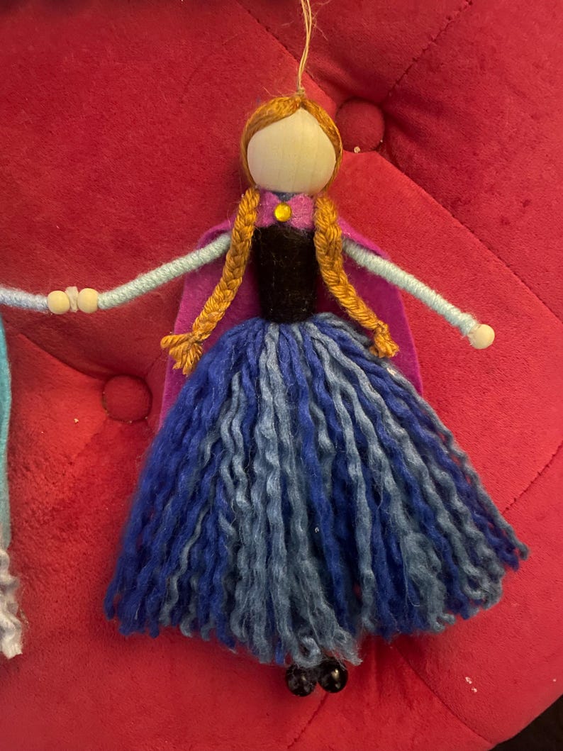 May include: A handmade felt doll with a wooden head, brown hair, and a blue and purple dress. The doll is wearing a purple cape and has a yellow button on her dress.