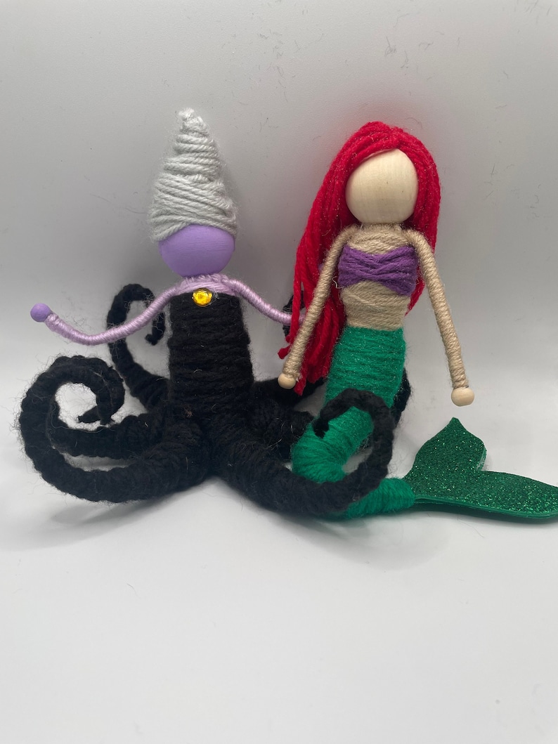 May include: Yarn doll figures of Ursula from The Little Mermaid and Ariel. Ursula is a black and purple yarn doll with a white yarn crown and tentacles. Ariel is a red yarn doll with a green yarn tail and a purple yarn top.