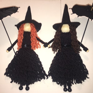 May include: Two yarn doll witches with black yarn dresses, black hats, and black umbrellas. One witch has orange yarn hair and the other has brown yarn hair.