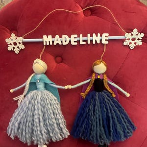 May include: A wooden sign with the name "MADELINE" hanging from a string with two felt dolls hanging below. The dolls are dressed as Elsa and Anna from the movie Frozen. The dolls are holding hands and have long yarn hair.