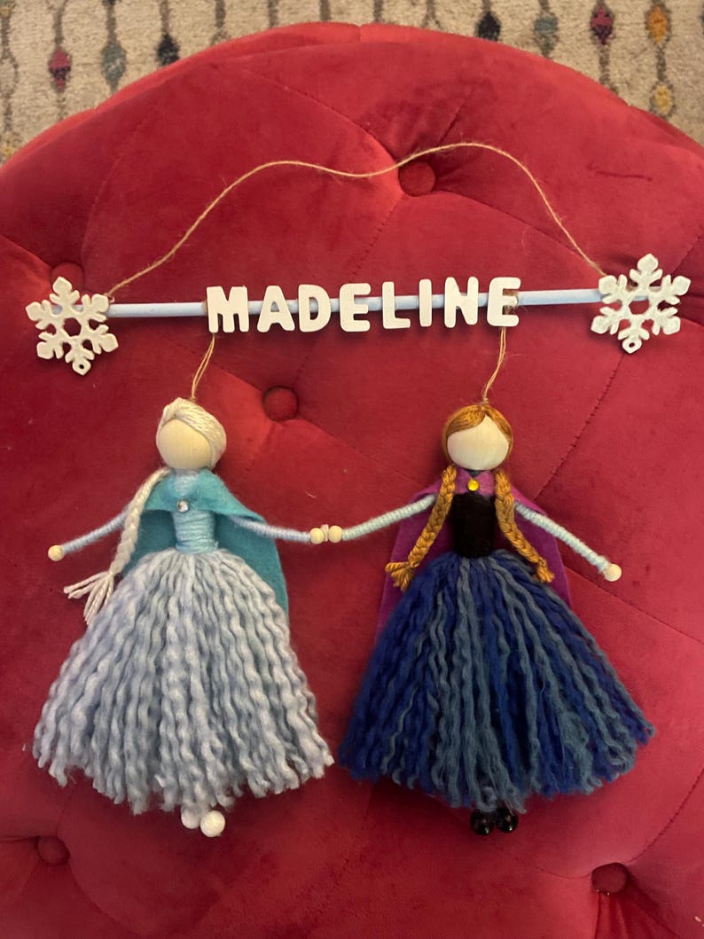 May include: A wooden sign with the name "MADELINE" hanging from a string with two felt dolls hanging below. The dolls are dressed as Elsa and Anna from the movie Frozen. The dolls are holding hands and have long yarn hair.