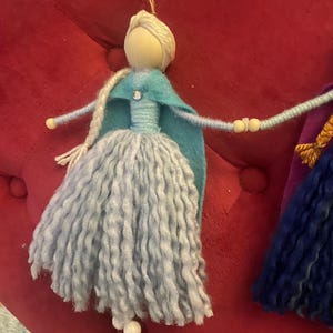 May include: A handmade doll with long, light blue yarn hair, a blue felt cape, and a light blue dress. The doll is holding hands with another doll, who is wearing a purple dress and a yellow crown. The dolls are hanging from a wooden sign that says "MADE WITH LOVE".