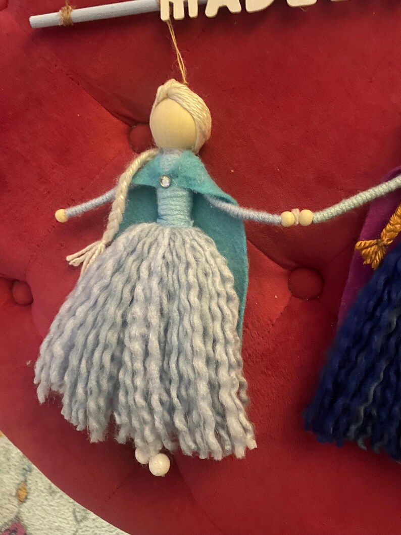 May include: A handmade doll with long, light blue yarn hair, a blue felt cape, and a light blue dress. The doll is holding hands with another doll, who is wearing a purple dress and a yellow crown. The dolls are hanging from a wooden sign that says "MADE WITH LOVE".
