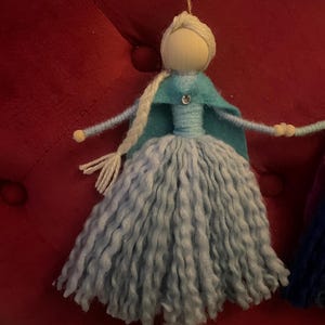 May include: A handmade yarn doll with a light blue dress, a teal cape, and a white yarn braid. The doll has a wooden head with a painted face and a sparkly blue gem on the cape.
