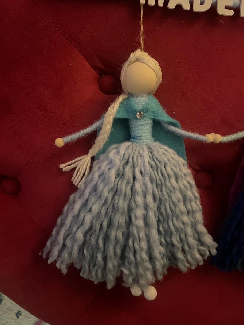 May include: A handmade yarn doll with a light blue dress, a teal cape, and a white yarn braid. The doll has a wooden head with a painted face and a sparkly blue gem on the cape.