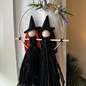 May include: Two black yarn witch dolls with red and brown yarn hair hanging from a gold metal hoop with a sprig of lavender and white flowers.