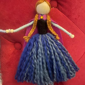 May include: A handmade doll with a wooden head, brown yarn hair, and a blue and purple yarn dress. The doll is wearing a purple felt cape and a black felt bodice with a yellow gem.