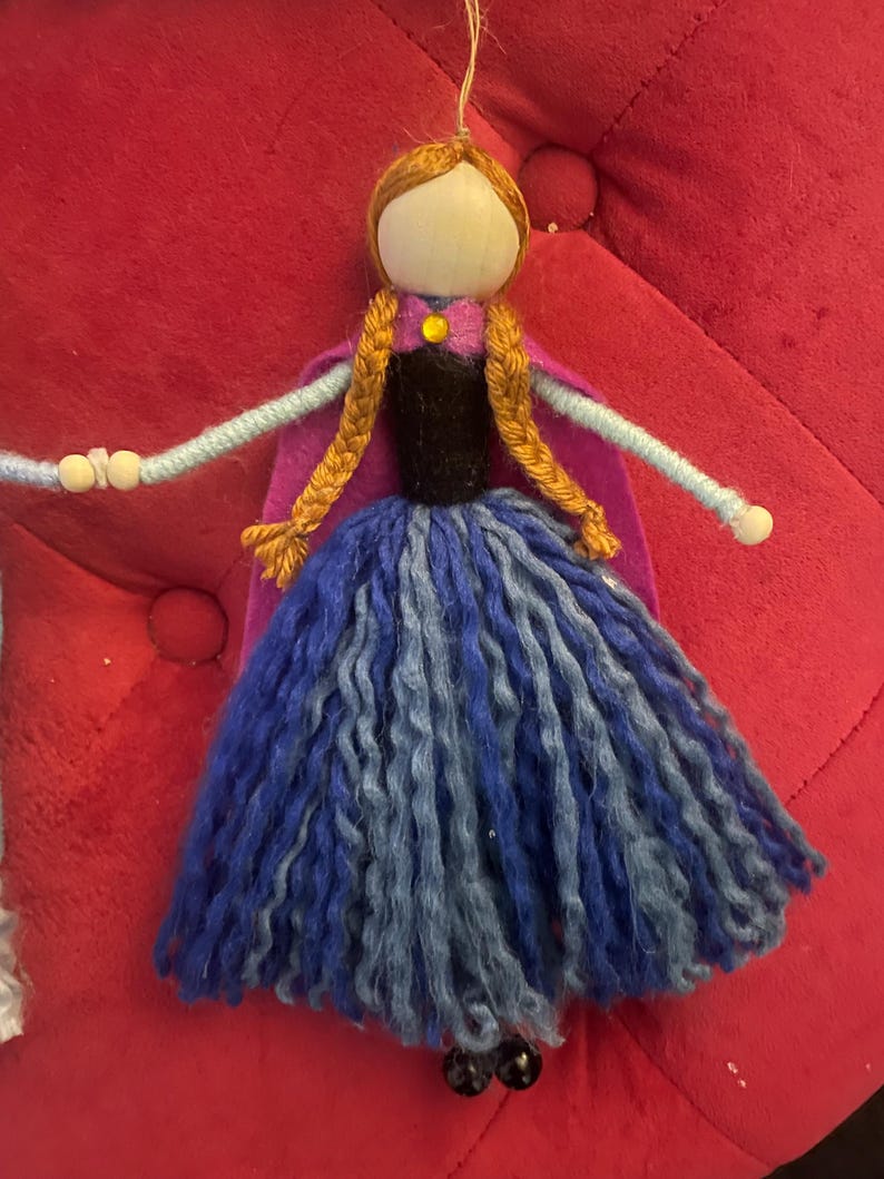 May include: A handmade doll with a wooden head, brown yarn hair, and a blue and purple yarn dress. The doll is wearing a purple felt cape and a black felt bodice with a yellow gem.