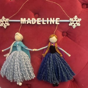 May include: A handmade felt mobile with two dolls dressed as Elsa and Anna from the movie Frozen. The dolls are hanging from a blue wooden dowel with the name "MADELINE" written in white letters. The mobile is decorated with two white snowflakes.