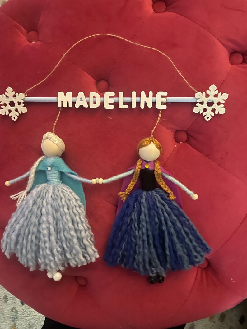 May include: A handmade felt mobile with two dolls dressed as Elsa and Anna from the movie Frozen. The dolls are hanging from a blue wooden dowel with the name "MADELINE" written in white letters. The mobile is decorated with two white snowflakes.