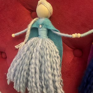 May include: A handmade doll with a blue and teal dress and a long, light blue yarn skirt. The doll has a wooden head with blonde yarn hair and a blue felt cape.