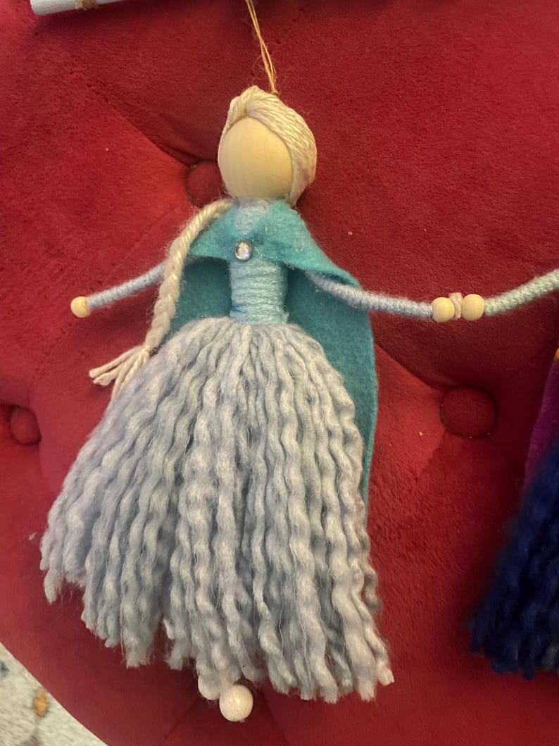 May include: A handmade doll with a blue and teal dress and a long, light blue yarn skirt. The doll has a wooden head with blonde yarn hair and a blue felt cape.