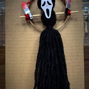 May include: A black yarn tassel with a gold ring and a white ghost face mask with a red knife on each side. The mask has a wide open mouth with a white scream.