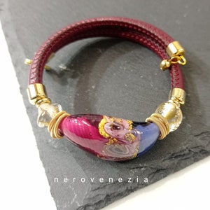Può includere: Un bracciale in pelle bordeaux con dettagli dorati e una perla centrale in vetro. La perla di vetro presenta sfumature di rosa, blu e oro. Un accessorio elegante.