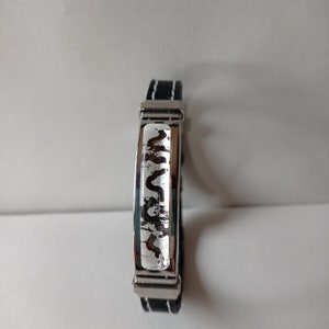 Könnte beinhalten: Ein schwarzes Lederarmband mit einem silbernen Band, das mit einem weißen abstrakten Design verziert ist.