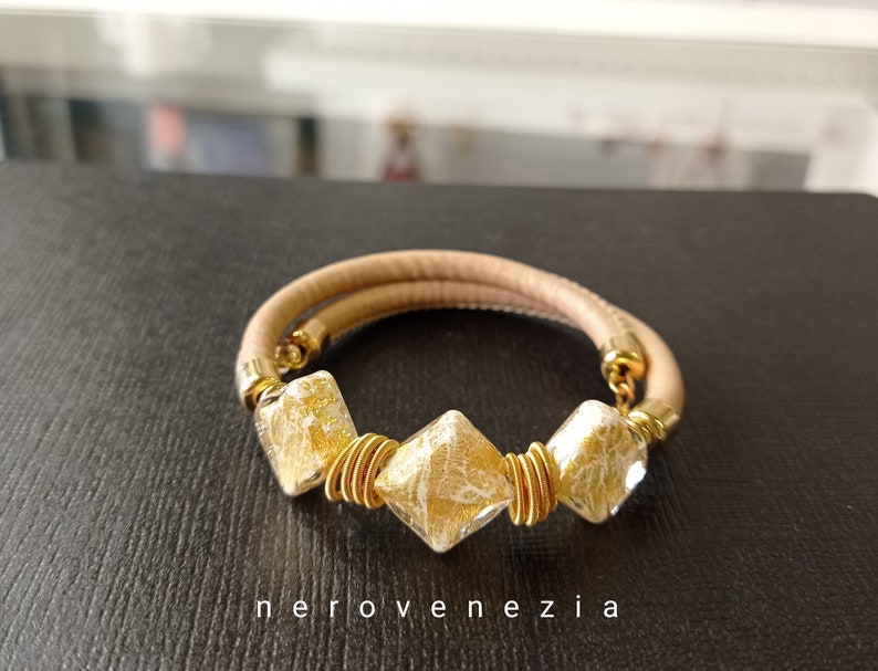 May include: A beige leather bracelet with three gold-toned square beads. The beads have a white, crackled glass design.