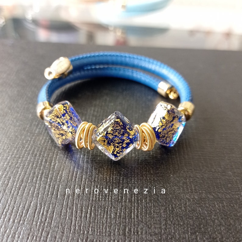 May include: A blue leather bracelet with three gold-wrapped square glass beads. The beads are filled with gold flakes.