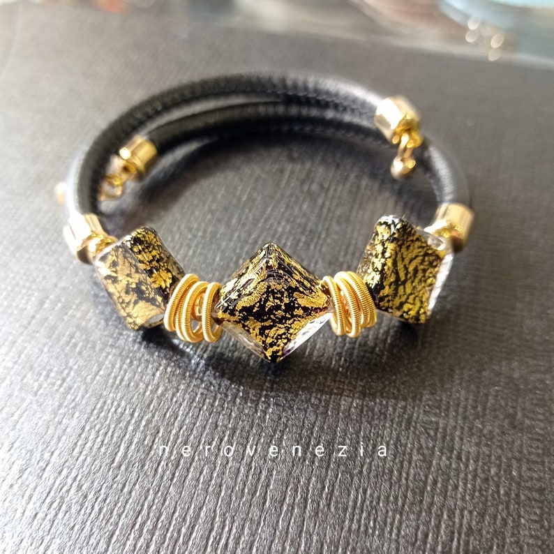 May include: A black leather bracelet with three gold-toned square glass beads. The beads have a black and gold speckled pattern.