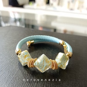 May include: A light blue leather bracelet with three gold-wrapped, square-shaped glass beads with gold flecks. The beads are arranged in a row on the bracelet.
