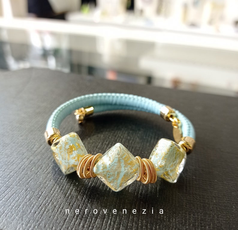 May include: A light blue leather bracelet with three gold-wrapped, square-shaped glass beads with gold flecks. The beads are arranged in a row on the bracelet.