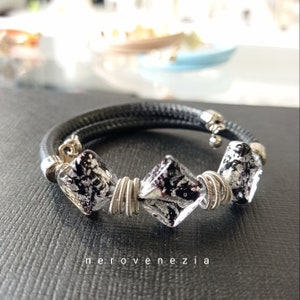 May include: Black leather bracelet with three silver and black square glass beads wrapped in silver wire. The bracelet has a silver charm attached to it.