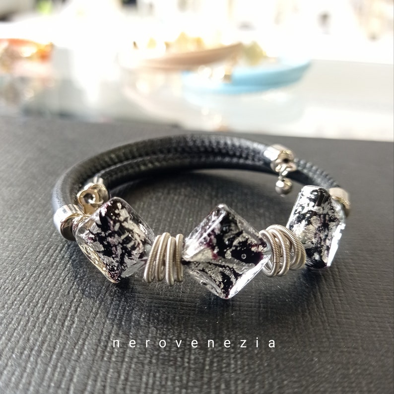 May include: Black leather bracelet with three silver and black square glass beads wrapped in silver wire. The bracelet has a silver charm attached to it.
