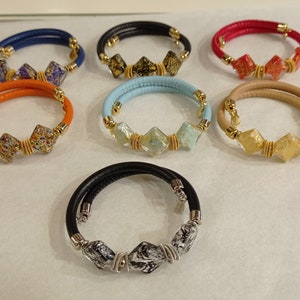 May include: Seven leather bracelets with different colored leather and glass beads. The beads are square shaped and have gold accents.