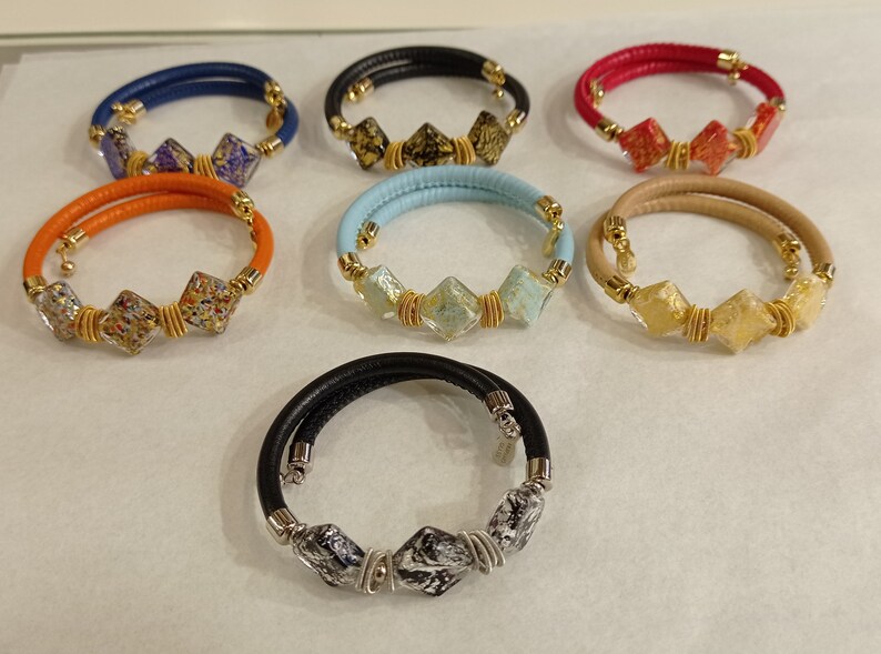 May include: Seven leather bracelets with different colored leather and glass beads. The beads are square shaped and have gold accents.