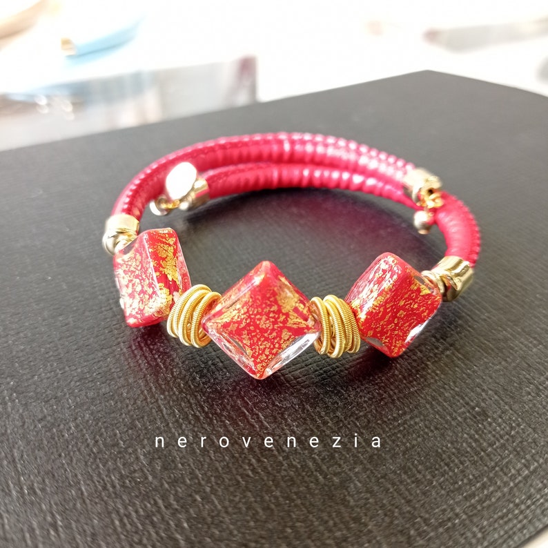 May include: Red leather bracelet with three gold-rimmed, red glass beads with gold flecks. The beads are square-shaped with rounded corners. The bracelet is wrapped twice around the wrist.