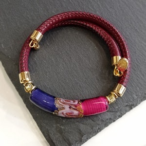 Può includere: Un braccialetto in pelle bordeaux con chiusura dorata e una perla di vetro multicolore. La perla è un mix di blu, rosa e bianco con pagliuzze dorate.