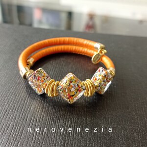 May include: A gold bracelet with three square-shaped glass beads. The beads are filled with colorful confetti and gold flakes. The bracelet is made of orange leather.
