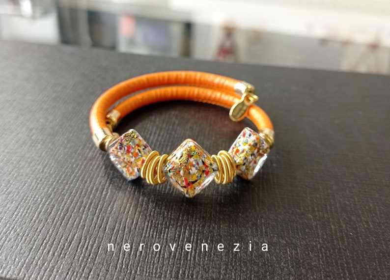 May include: A gold bracelet with three square-shaped glass beads. The beads are filled with colorful confetti and gold flakes. The bracelet is made of orange leather.