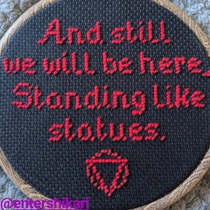 May include: A black embroidery hoop with red embroidery that reads "And still we will be here Standing like statues."