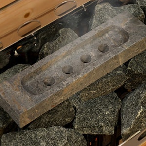 May include: A grey stone sauna heater with five holes for pouring water. The heater is surrounded by grey and green rocks.