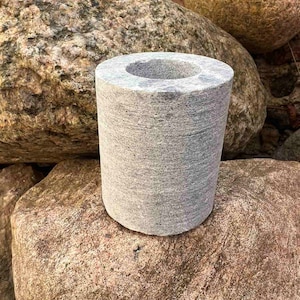 Può includere: Un vaso cilindrico in pietra grigia con una superficie liscia e testurizzata. Il vaso è appoggiato su una grande roccia grigia.