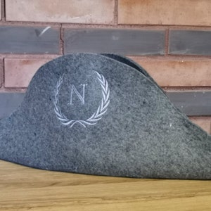 Puede incluir: Un sombrero de tres picos de fieltro gris con una corona de laurel bordada en blanco y la letra "N" en el centro.