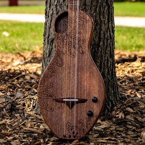 May include: A wooden acoustic guitar with intricate floral carvings on the body. The guitar is leaning against a tree trunk in a grassy field.
