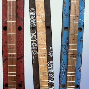 May include: Three wooden cigar box guitars in their cases. The cases are decorated with different patterns. The guitars are all different colors: red, brown, and blue.