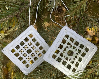 Op de afbeelding: Twee witte, vierkante, gehaakte ornamenten met opengewerkte ontwerpen, hangend aan witte touwtjes tegen een achtergrond van groene dennennaalden. De ornamenten hebben ingewikkelde patronen.