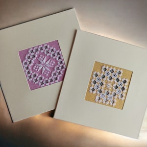 May include: Two white cards with embroidered designs. The card on the left has a pink background with a white snowflake design. The card on the right has a yellow background with a white geometric design.