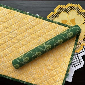 May include: A yellow quilted table runner with a green floral border. The runner is rolled up and has a green floral fabric underneath.