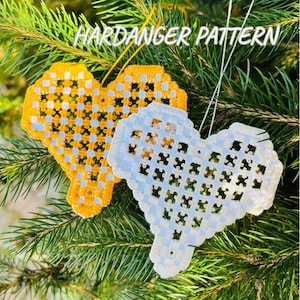 Peut inclure: Deux ornements en forme de cœur avec un motif Hardanger, un jaune et un blanc, sont suspendus à un sapin de Noël. L'ornement jaune a un motif tissé bleu et noir, tandis que l'ornement blanc a un motif tissé bleu et orange. Le texte "HARDANGER PATTERN" est en haut.