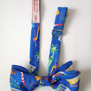 May include: A blue bow tie featuring a colorful school supply pattern including crayons, calculators, and paint splatters. The fabric strap includes a metal adjustable slider and a fabric label that reads Hand Crafted by About Bow Ties.