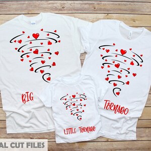 Puede incluir: Tres camisetas blancas con un diseño de tornado de corazones rojos. Las camisetas tienen las palabras "BIG", "LITTLE TORNADO" y "TORNADO" en rojo. Sobre un fondo de madera.