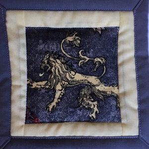 May include: A gray and yellow fabric coaster with a black and white lion design. The lion is depicted with a crown and a tail.