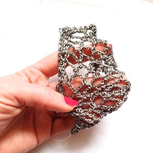 以下が含まれることがあります： 白い背景に、銀色のオープンウィーブのブレスレットが写っています。 ブレスレットは、幾何学模様の繊細なチェーンのような構造をしています。 ブレスレットを持っている手にはピンクのマニキュアが塗られています。