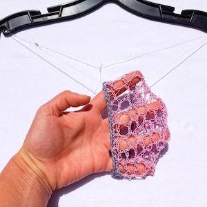 Pode incluir: Uma máscara facial de crochê, cor lavanda, com uma borda cinza, é segurada por uma pessoa. A máscara está suspensa em um cabide preto com fio branco.
